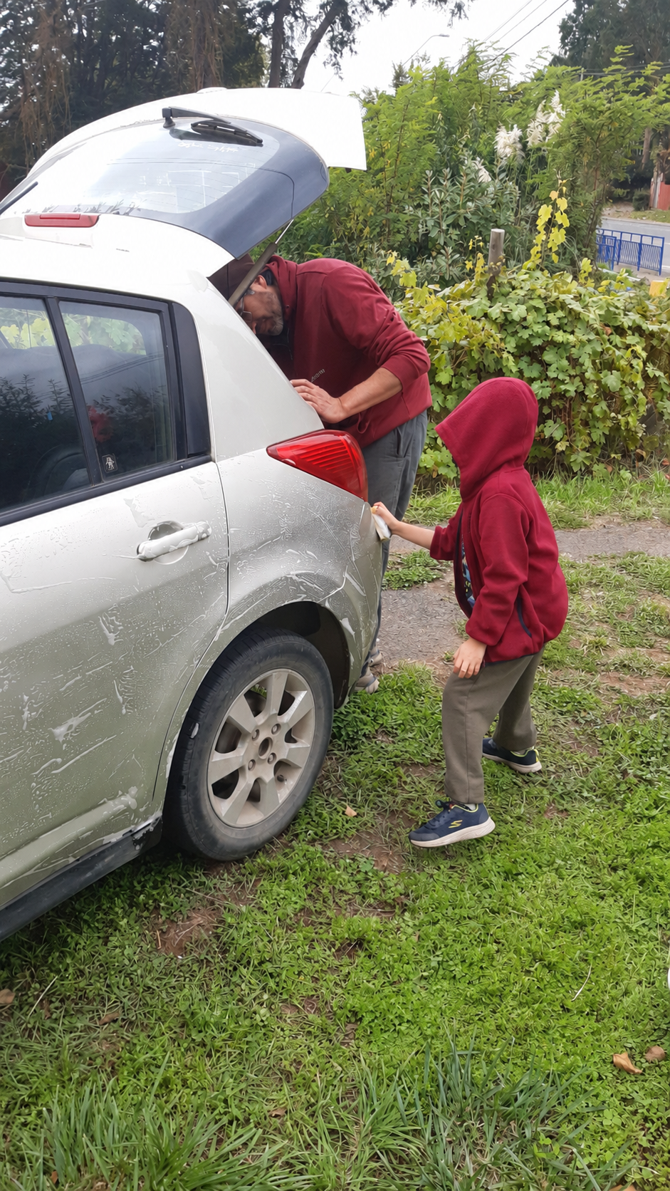 Un niño con el rostro cubierto por su capucha ayuda a su papá a lavar el auto